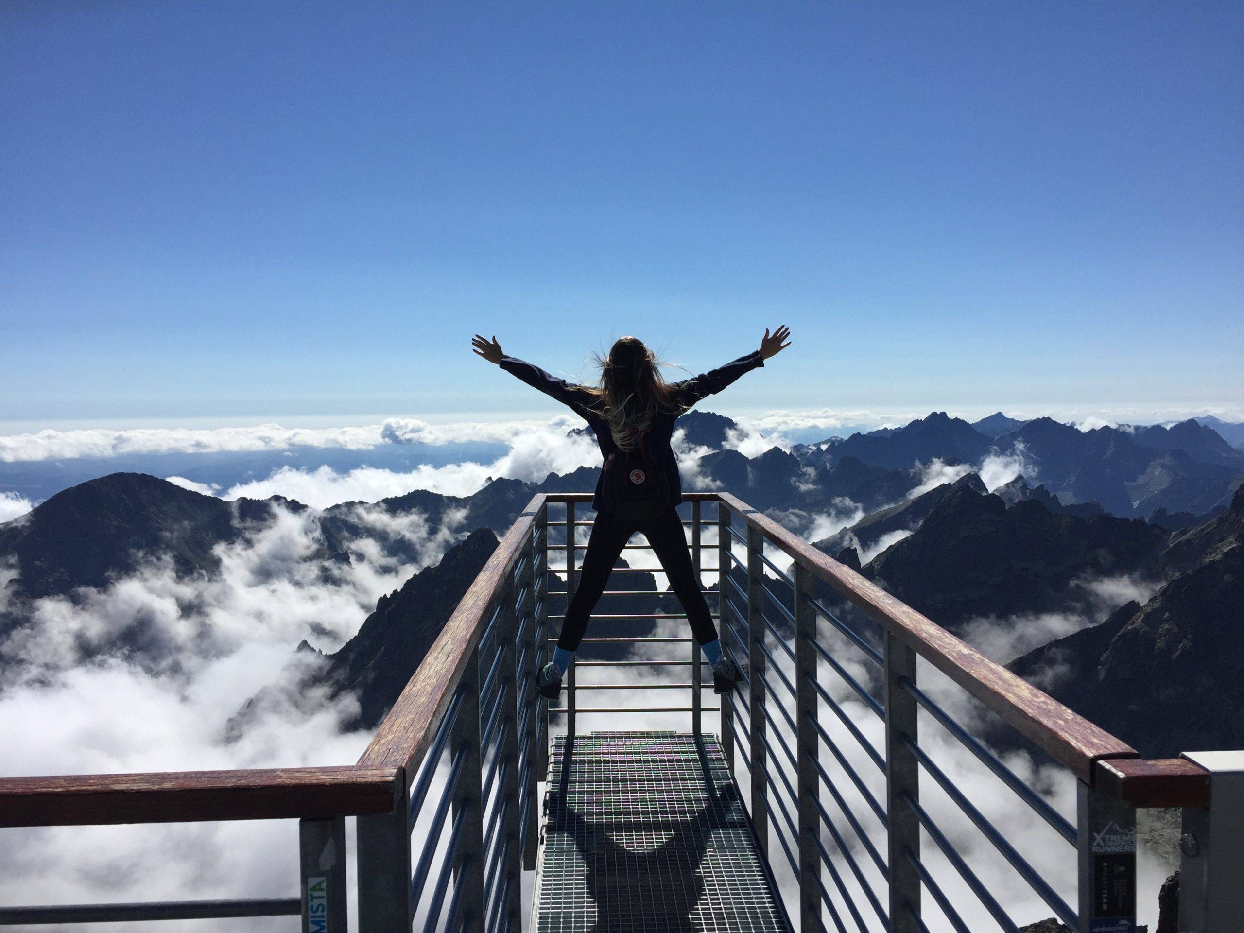 Person standing on hand rails facing cloudy mountaintops